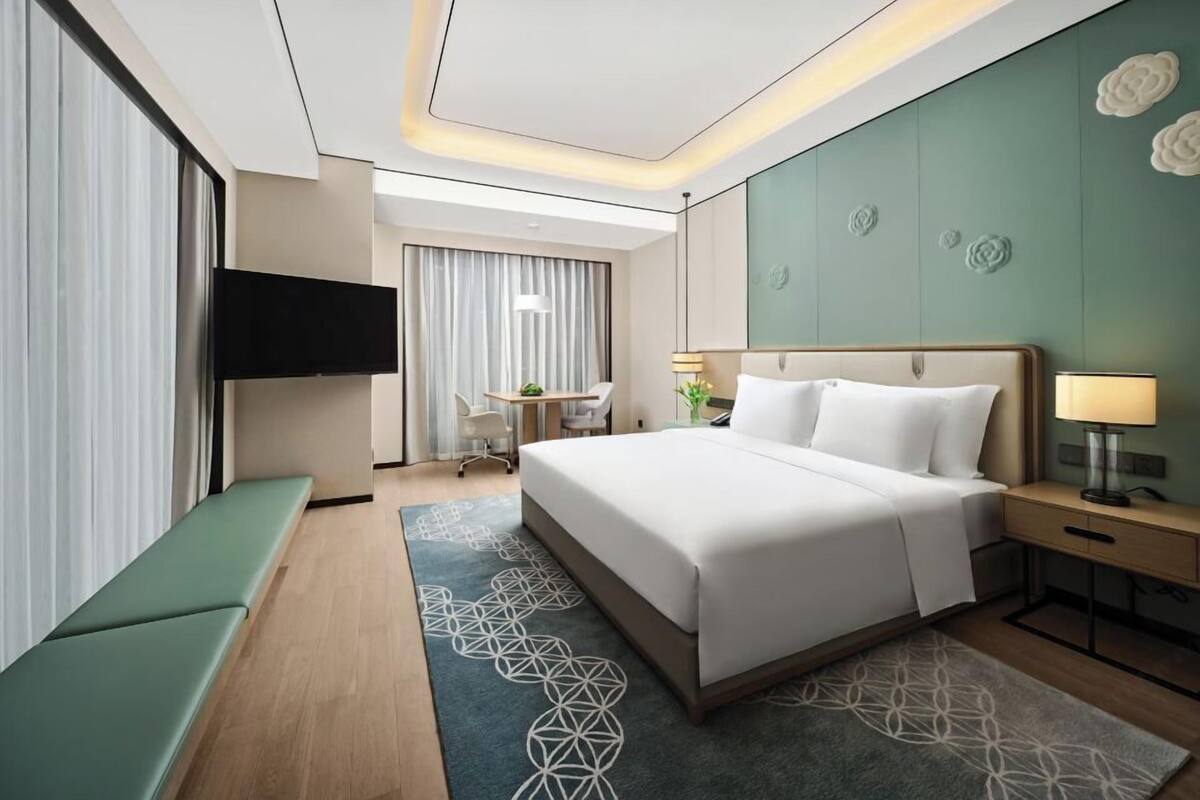 A modern hotel room with a large bed, a desk, a TV, and a seating area.