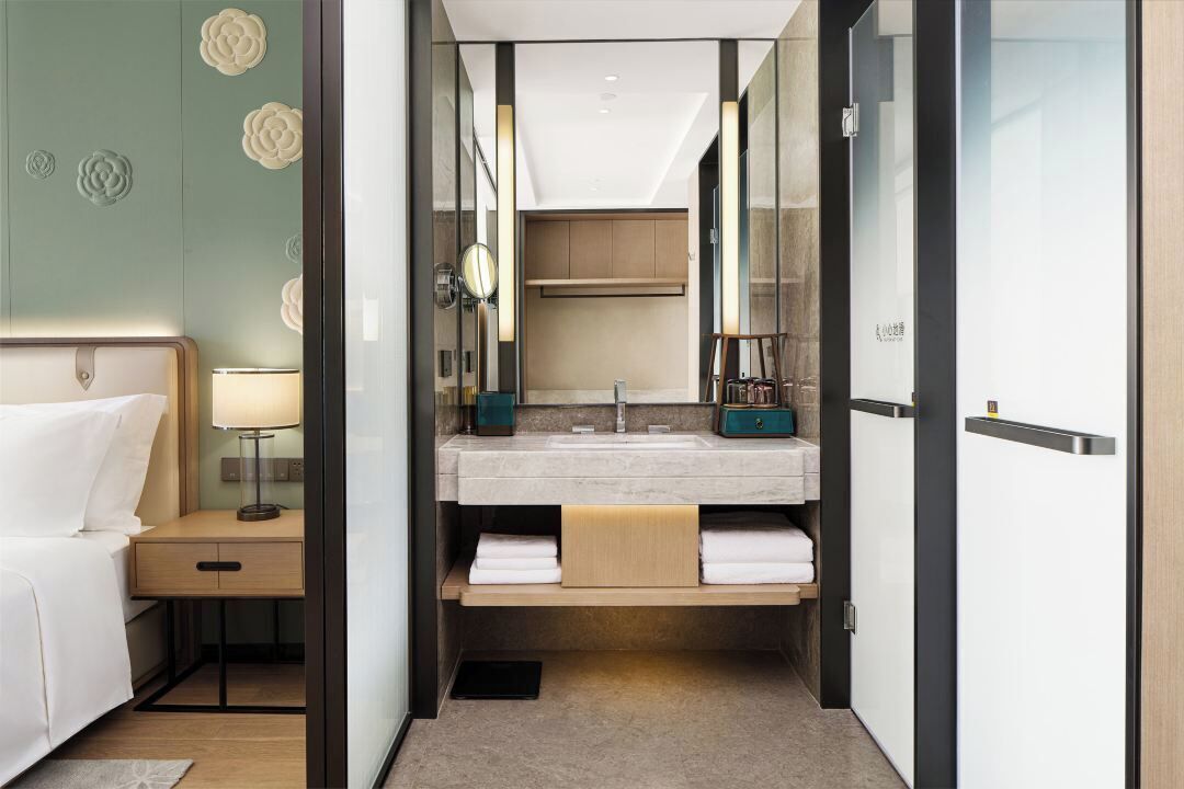 A modern hotel room with a bed, bedside table, lamp, and a bathroom with a sink, mirror, and towel storage.