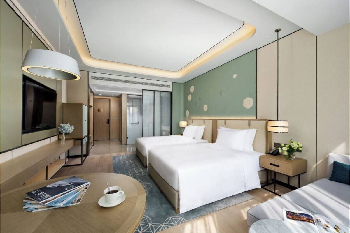 A modern hotel room with two beds, a TV, a desk with a chair, and a dining area.