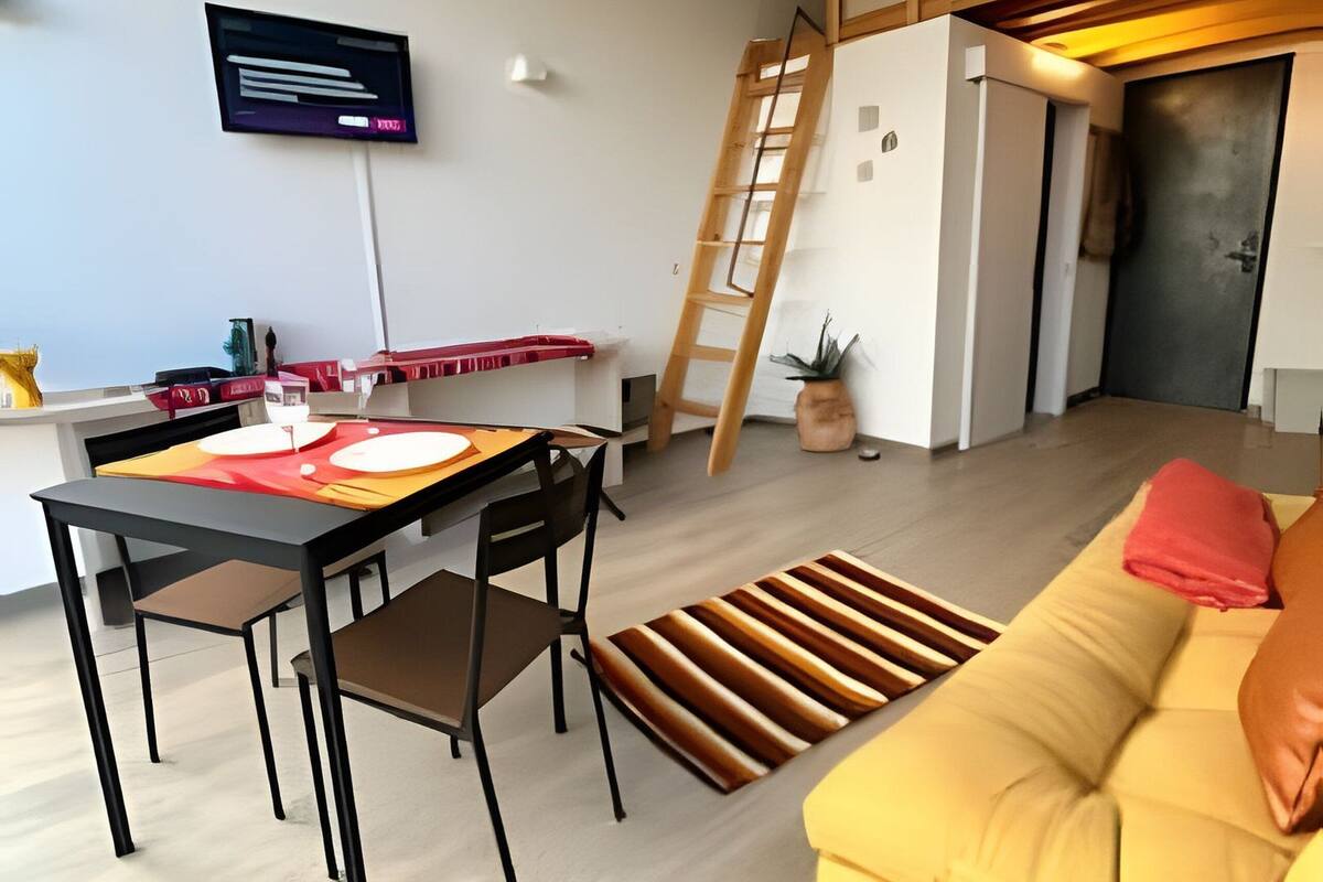 A modern living space with a dining table, chairs, a sofa, a TV mounted on the wall, a ladder, and a door.