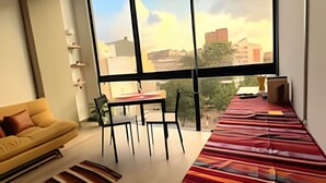 A modern living room with a large window, a yellow sofa, a dining table with chairs, and a colorful rug. - Tu hogar en Chapinero alto te espera (Bogotá)