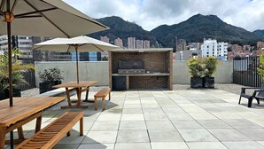 An outdoor patio with a brick barbecue, wooden tables and benches, and a TV screen. - Tu hogar en Chapinero alto te espera (Bogotá)