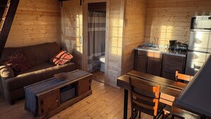 A cozy cabin interior with a wooden floor, a sofa, a bench, a dining table with chairs, a kitchen area with a sink and appliances, and a window with curtains. - Shawnee Wine Trail Farm Stay (Makanda)