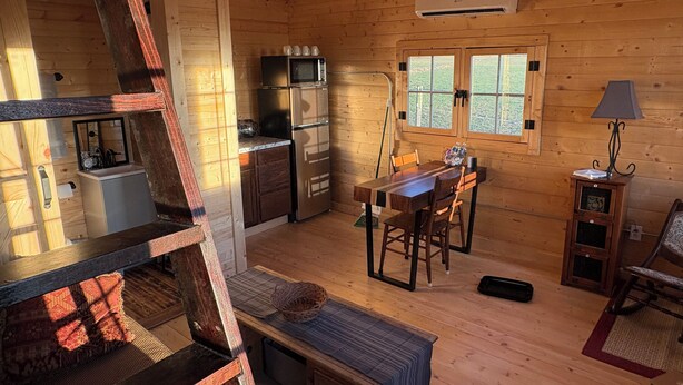 A cozy cabin interior with a wooden interior, a dining area with a table and chairs, a kitchen with a refrigerator and microwave, and a window with a view of greenery. - Shawnee Wine Trail Farm Stay (Makanda)