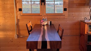 A wooden cabin interior with a dining table, chairs, a window with a view of the outdoors, and an air conditioning unit above the window. - Shawnee Wine Trail Farm Stay (Makanda)