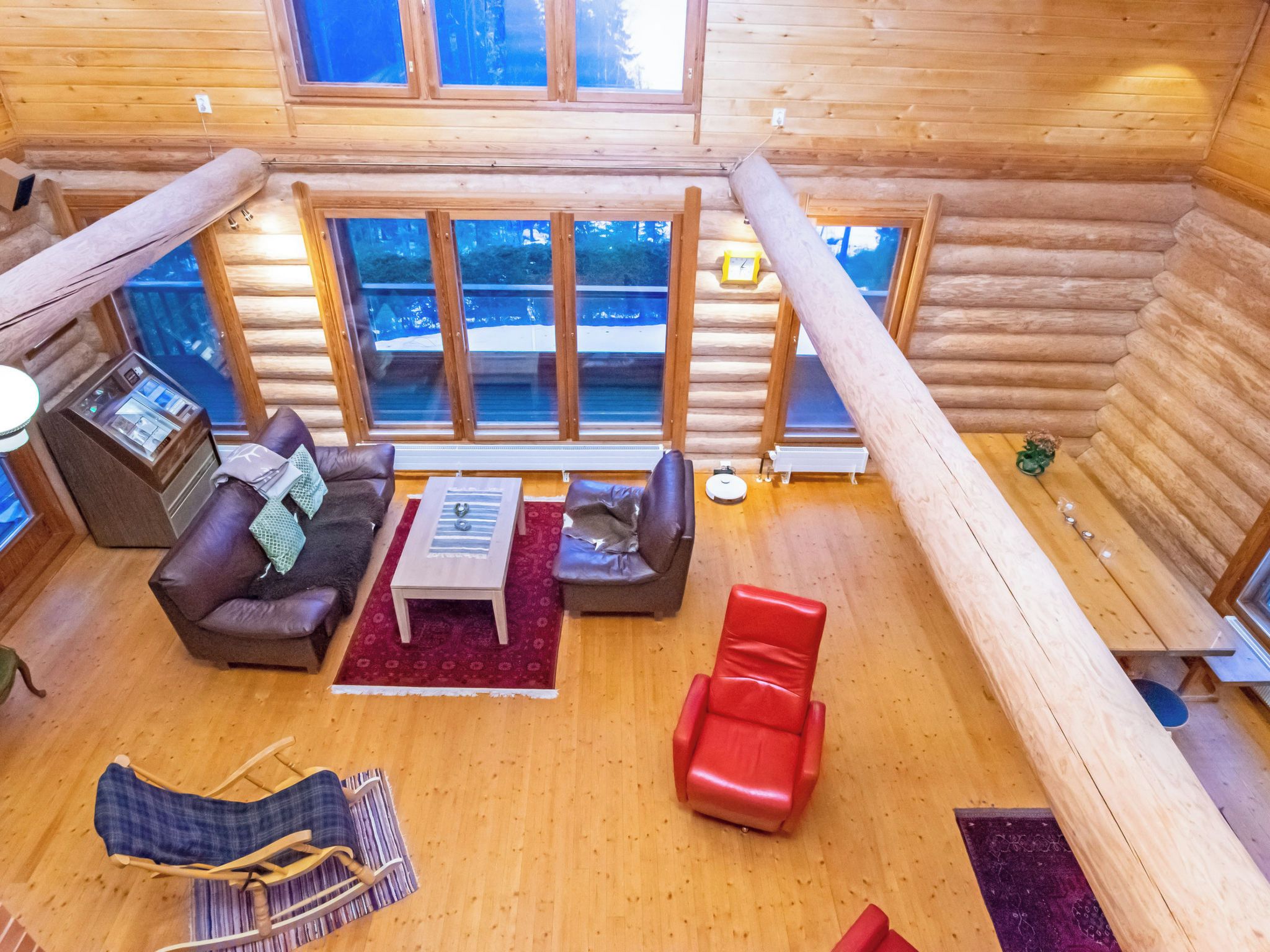 Hirsihirrestä rakennetun mökin sisätilat, joissa on oleskelutila, jossa on sohvaryhmä, punainen tuoli, pieni pöytä ja ikkuna, josta avautuu näkymä lumiseen maisemaan.