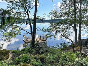 Une terrasse en bois, dotée de rampes de protection, qui offre une vue sur un lac paisible, le tout entouré d’arbres et de verdure.