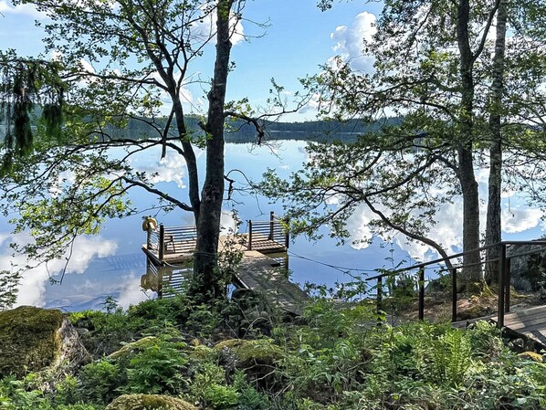 A wooden deck with railings overlooking a calm lake, surrounded by trees and greenery.