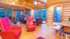 A log cabin interior with a wooden ceiling, large windows, and a seating area with red and black chairs.