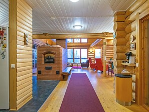 L’intérieur d’une cabane en rondins, avec une cheminée, des murs en bois, un coin repas et une vue sur l’extérieur grâce à de grandes fenêtres.