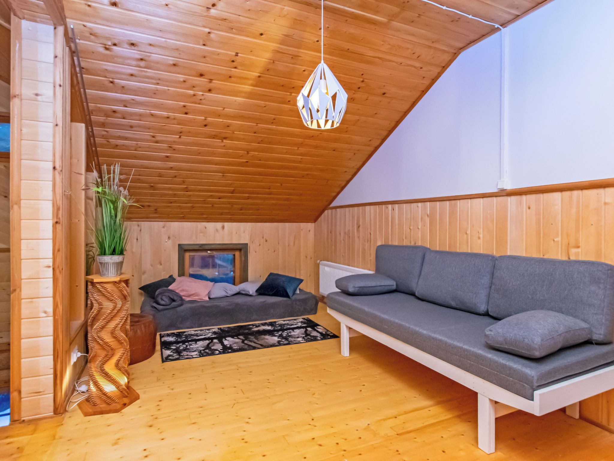 Tunnelmallinen huone, jossa on puinen katto, sohva, tyynyillä varustettu sänky, ruukkukasvi ja riippuvalaisin.
