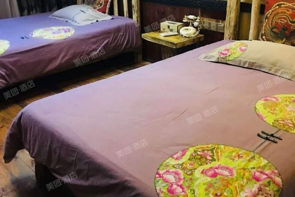 Habitación con dos camas, cada una con colcha morada y almohada estampada. Una cama tiene cabecera de madera, y la otra, una mesita de noche de madera con lámpara y teléfono.