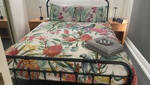 Un lit avec une couvre-lit à motifs floraux et un oreiller assorti, une table de chevet avec une lampe et une petite table de nuit avec des livres.