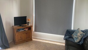 Une pièce avec un téléviseur posé sur un meuble en bois, une fenêtre avec un store et un canapé avec un coussin.