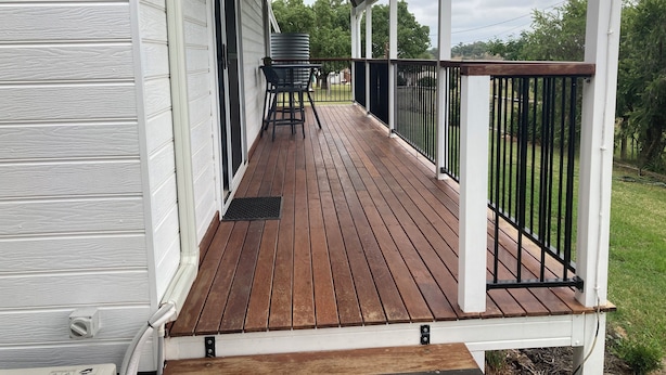 Une terrasse en bois couverte, dotée d’une rambarde en métal, d’une table et de chaises, et offrant une vue sur une cour verdoyante.