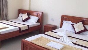 A hotel room with two wooden beds, each with a white mattress and matching pillows. There is a small wooden nightstand between the beds, and a wall-mounted lamp above the right bed.