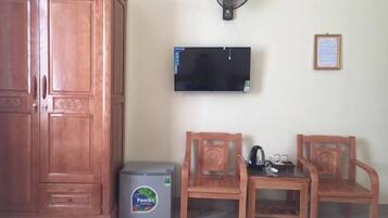 A room with a wooden wardrobe, a small refrigerator, two wooden chairs, a table, a wall-mounted TV, and a standing fan.