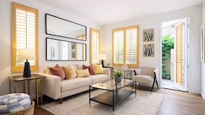 Un salon comprenant un canapé, des fauteuils, une table basse et un tapis. Il y a deux fenêtres avec des stores, une porte donnant sur l’extérieur et des œuvres d’art accrochées aux murs.