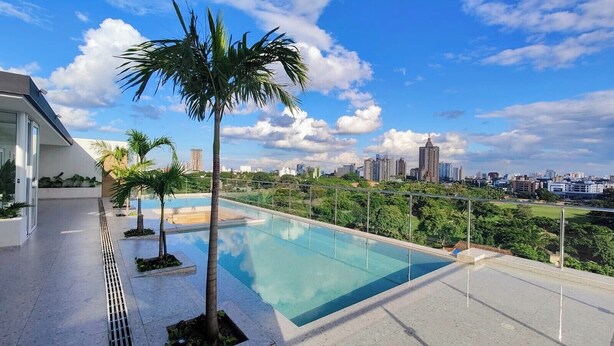 Una piscina en la azotea con agua cristalina, rodeada de palmeras y un paisaje urbano al fondo.