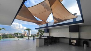 Una cocina exterior moderna con vista a la piscina, que incluye una zona de bar, una parrilla empotrada y amplias velas de sombra.