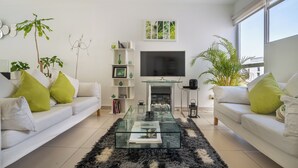 Una sala de estar moderna con una mesa de centro de vidrio, sofás blancos con cojines verdes, una chimenea y un ventanal grande.