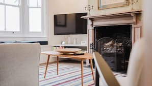 Ein Wohnzimmer mit Kamin, Flachbildfernseher, Tisch mit Schüssel und Fernbedienung sowie Teppich mit Streifen.
