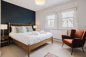 A bedroom with a wooden bed, two bedside tables with lamps, a brown armchair, and a window with blinds. - Stunning 3 Bedroom Oxford Retreat (Oxford)