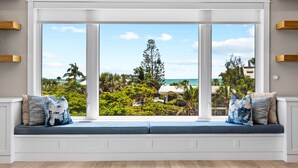 Een kamer met een groot raam met uitzicht op een tropisch landschap, een ingebouwde bank met kussens en houten planken aan de wanden.