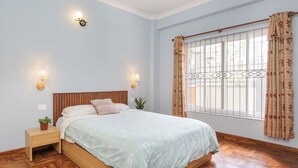 A bedroom with a wooden bed, two bedside lamps, a nightstand with a plant, and a window with curtains.
