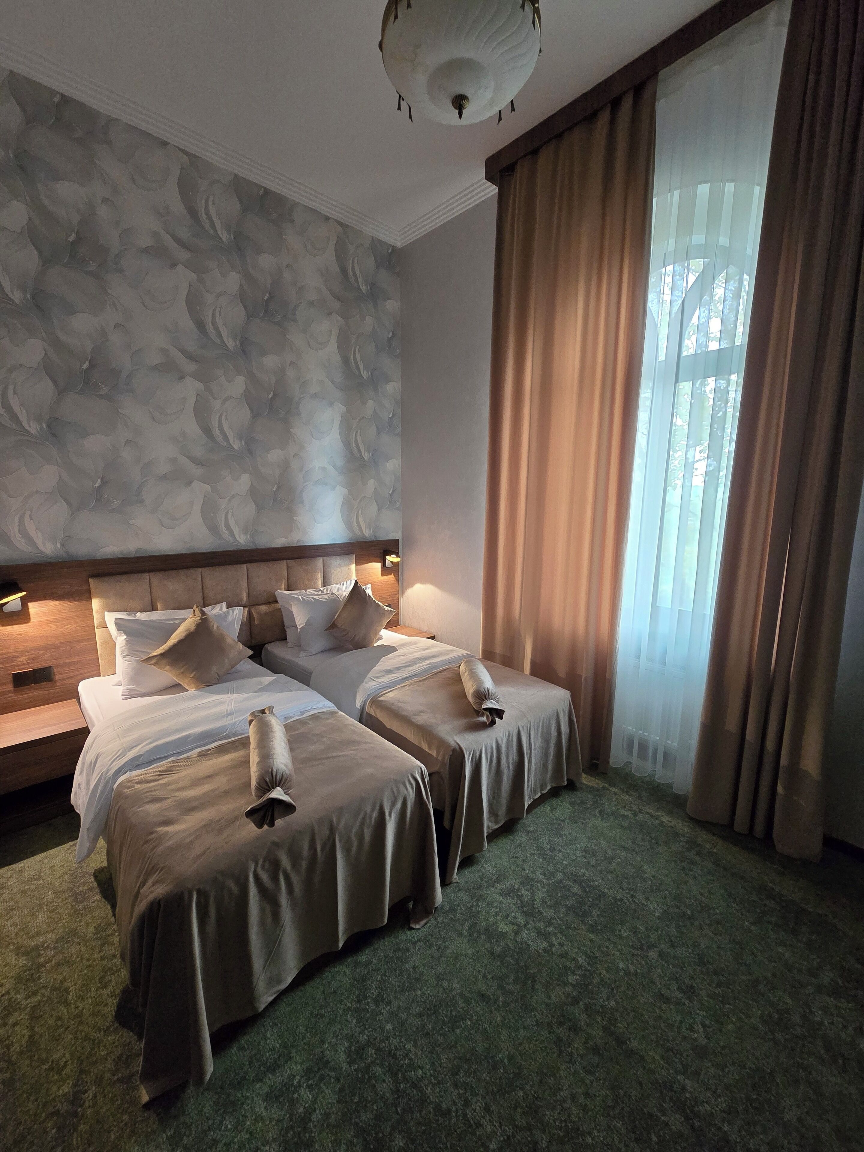 A hotel room with two beds, a large window with curtains, a patterned wall, and a ceiling light.