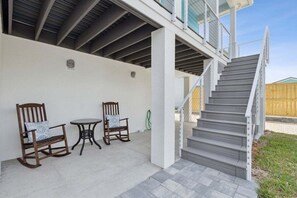 Un porche cubierto con dos mecedoras y una mesita, una escalera que lleva a un nivel superior y una valla de madera al fondo.