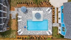Vue aérienne d’un bâtiment moderne doté d’une piscine rectangulaire, entourée de chaises longues blanches et de parasols, et jouxtant un bâtiment bleu.