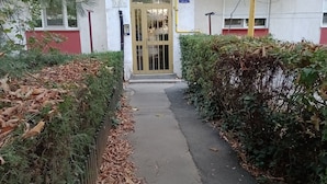Un chemin menant à un bâtiment dont l’entrée est une porte en verre, le tout étant entouré de verdure et de feuilles mortes.