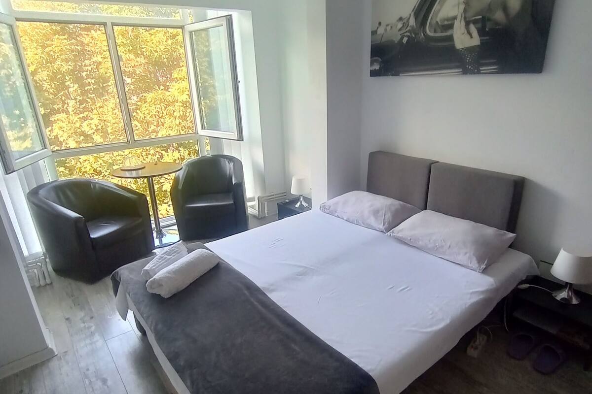 Un dormitorio con cama, una silla, una mesa, un ventanal y un cuadro.