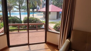 A room with a large sliding glass door leading to a pool area, a wooden deck, and a view of a building with a red roof. - Palmeraiebeach Resort Rayong (Klaeng)