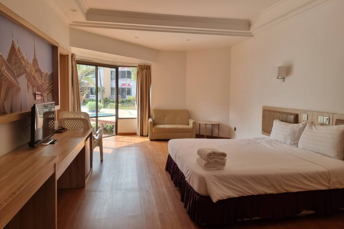A hotel room with a bed, a desk with a computer, a chair, and a view of a swimming pool and buildings.
