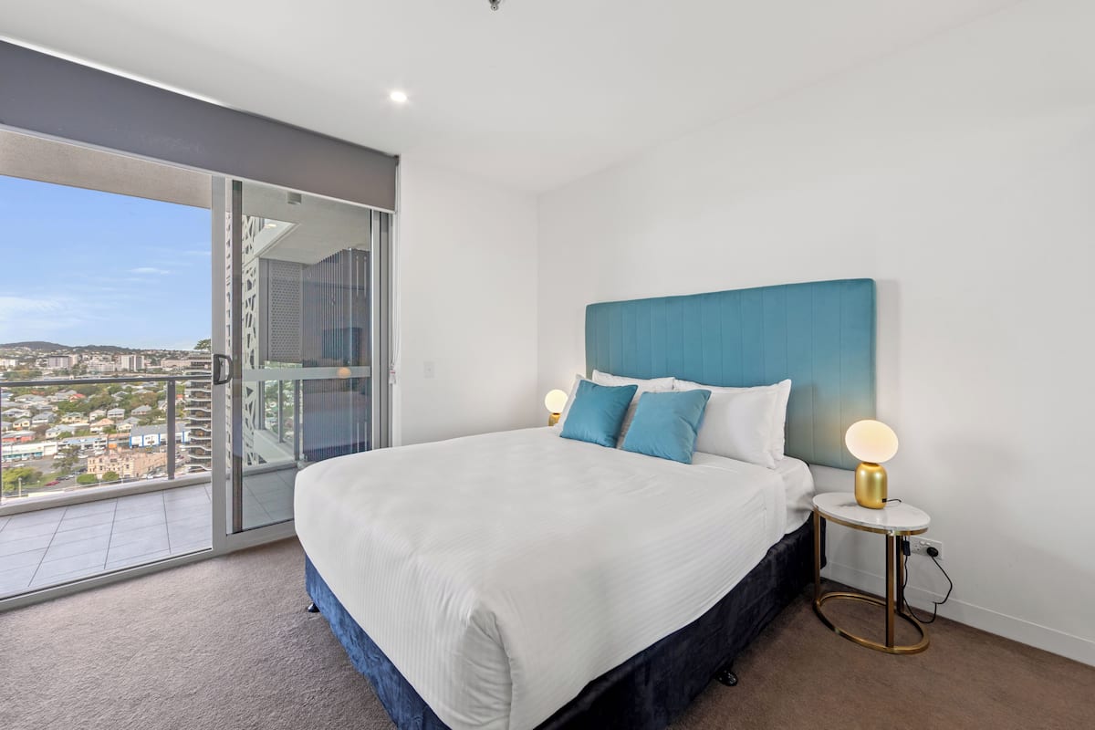 A modern bedroom with a large bed, a bedside table with a lamp, and a glass door leading to a balcony with a city view.