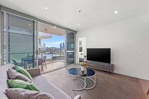 A modern living room with a sofa, a round coffee table, a flat-screen TV, and a sliding glass door leading to a balcony with a city view. - Trafalgar Lane Apartments by CLLIX (Woolloongabba)