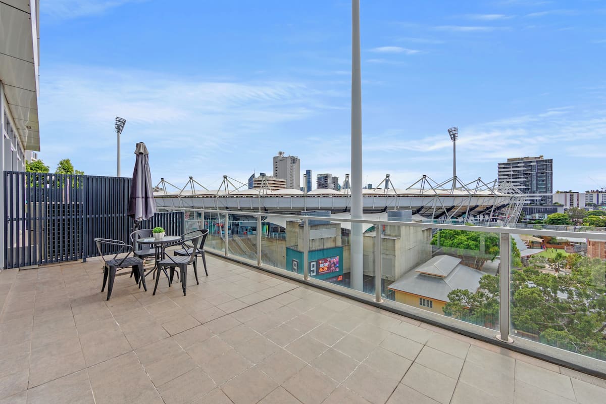 A balcony with a view of a stadium, a glass railing, and a seating area with chairs and a table.