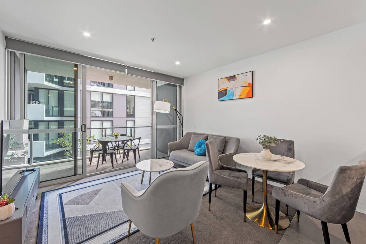 A modern living room with a sofa, two armchairs, a coffee table, and a dining table. There is a large window with a view of buildings outside.