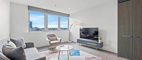 Un salon moderne avec un canapé, un fauteuil, un téléviseur et une grande fenêtre donnant sur la ville.