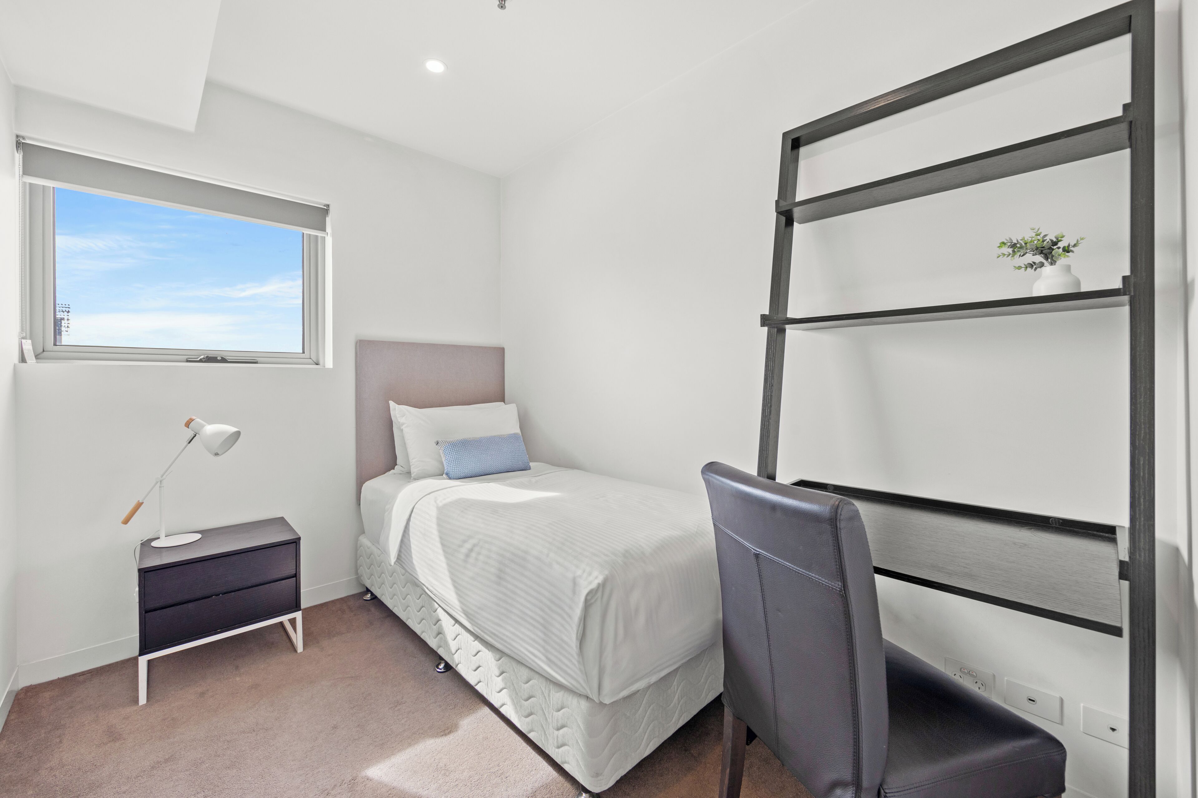 A bedroom with a bed, a desk, a chair, a nightstand, a lamp, and a window with a view of the sky.