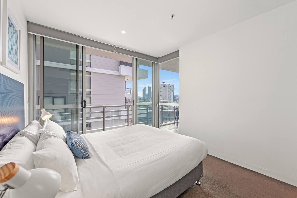 A modern bedroom with a large bed, a balcony with glass doors, and a city view.