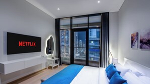 A modern hotel room with a large bed, a flat-screen TV mounted on the wall, a city view through floor-to-ceiling windows, and a minimalist design.