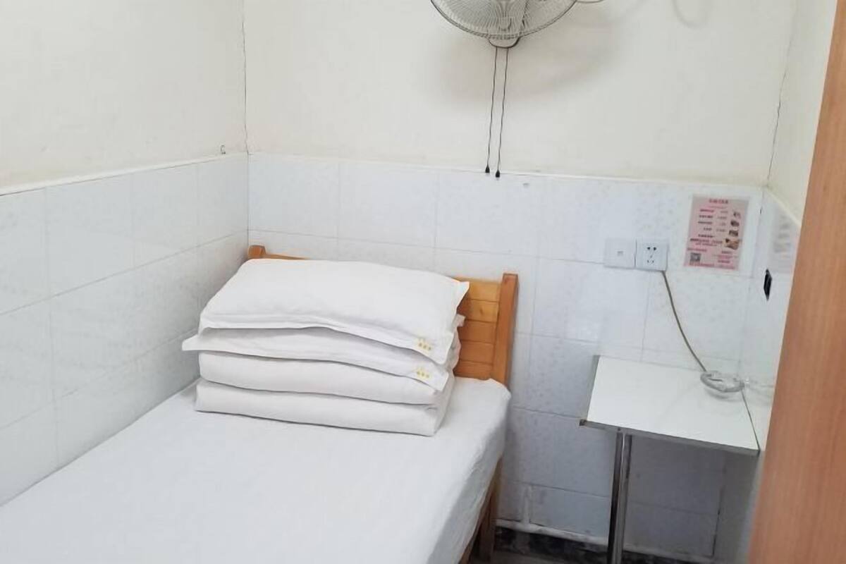 Une petite chambre avec un lit simple, une tête de lit en bois, un matelas blanc, une petite table de chevet et un ventilateur fixé au mur.