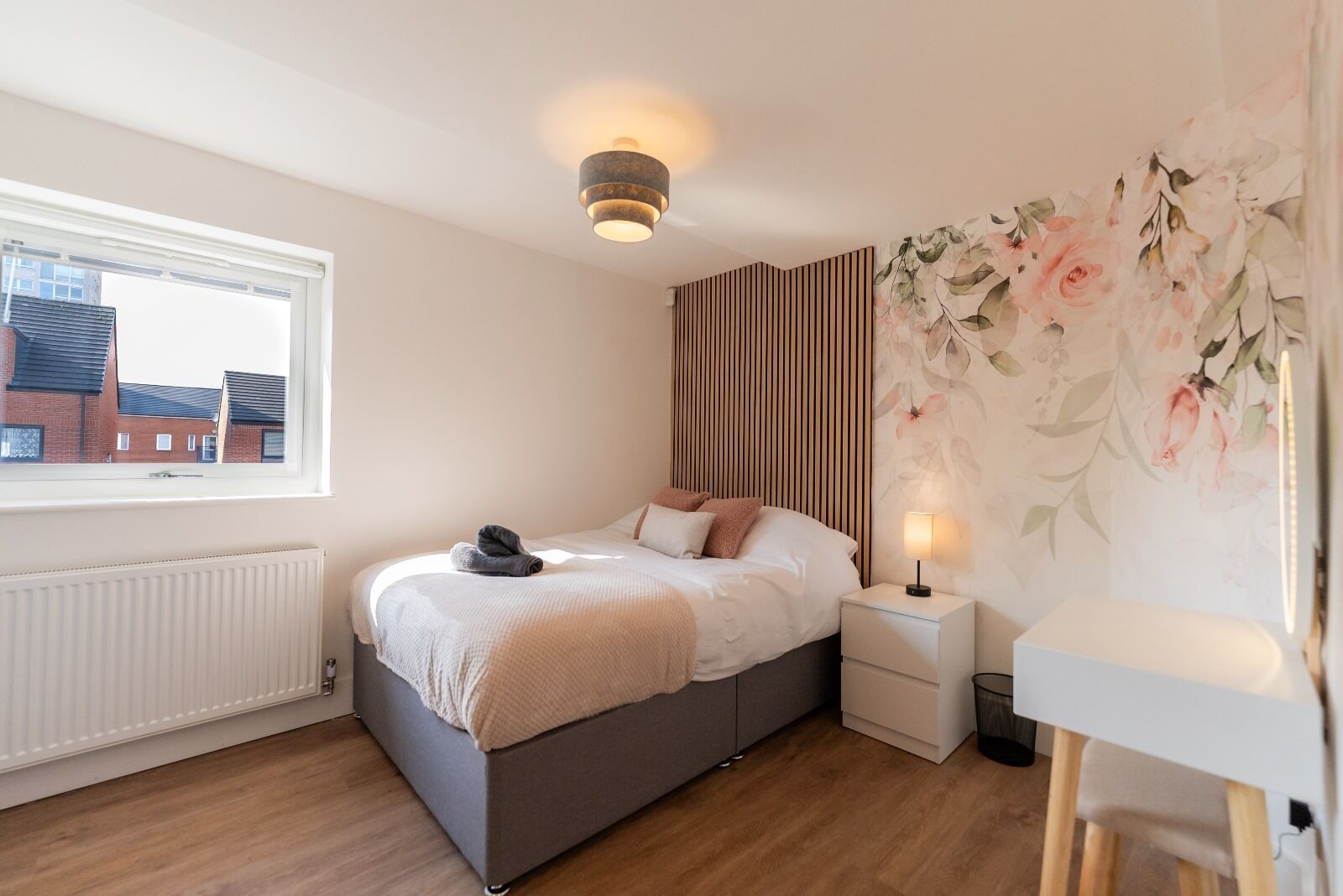 Une chambre à coucher comprenant un lit, une table de chevet, une lampe, un radiateur et une fenêtre donnant sur des immeubles.