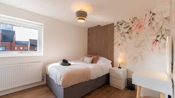Une chambre à coucher comprenant un lit, une table de chevet, une lampe, un radiateur et une fenêtre donnant sur des immeubles.