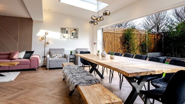 A modern living room with a large wooden dining table, black chairs, a sofa, and a bench. There are skylights and a view of the outdoors. - Luxury 8 Bed Home Manchester (Manchester)