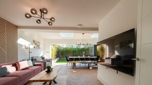 A modern living room with a pink sofa, a wooden coffee table, and a dining area with a table and chairs. There is a TV mounted on the wall and a ceiling light fixture. - Luxury 8 Bed Home Manchester (Manchester)