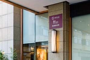 Ein Hotel Eingang mit Schild, einer modernen Leuchte und einer Glastür, die das Innere des Gebäudes reflektiert.